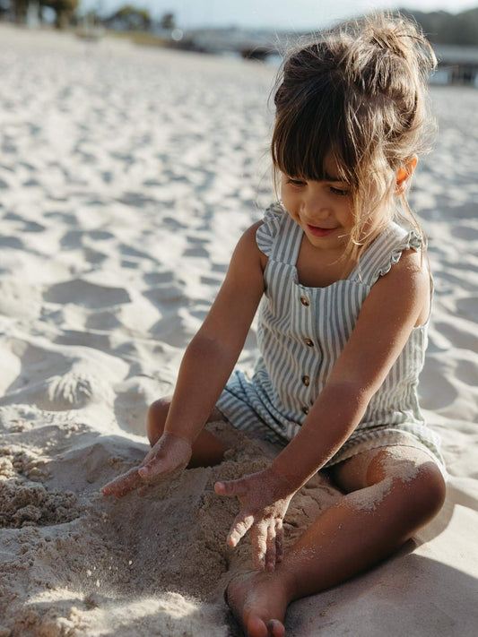 A young girl with her hair in a messy bun sits on a sandy beach, playing with sand while wearing a sleeveless, light blue and white striped romper with gold buttons.