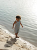 A young child wearing a blue and white striped sleeveless jumpsuit stands barefoot at the edge of a calm body of water on a sandy shore.