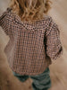 Child wearing a checkered shirt with ruffled shoulders in a natural setting Ruffle Woven Blouse - Muslin Cotton Check Girls