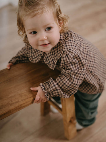 Child in a checkered shirt standing on a wooden stool indoors Ruffle Woven Blouse - Muslin Cotton Check Girls