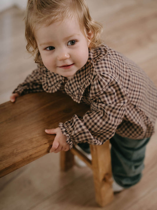 Child in a checkered shirt standing on a wooden stool indoors Ruffle Woven Blouse - Muslin Cotton Check Girls