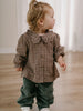 Child wearing a plaid shirt and green pants standing on a wooden floor. Ruffle Woven Blouse - Muslin Cotton Check Girls
