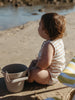 A toddler sits on a sandy beach next to a small bucket, wearing a sleeveless cream and tan striped romper. The background shows water and rocks in soft focus.