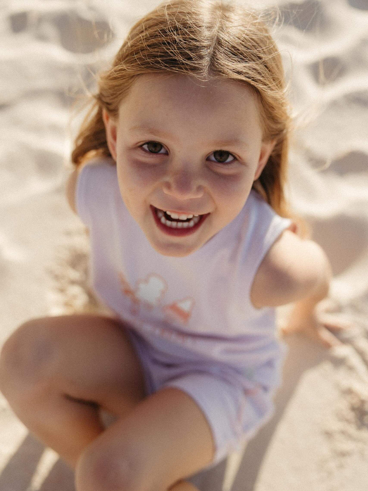 A young girl with light brown hair smiling at the camera while crouching in the sand, wearing a pale lavender sleeveless top and matching shorts.