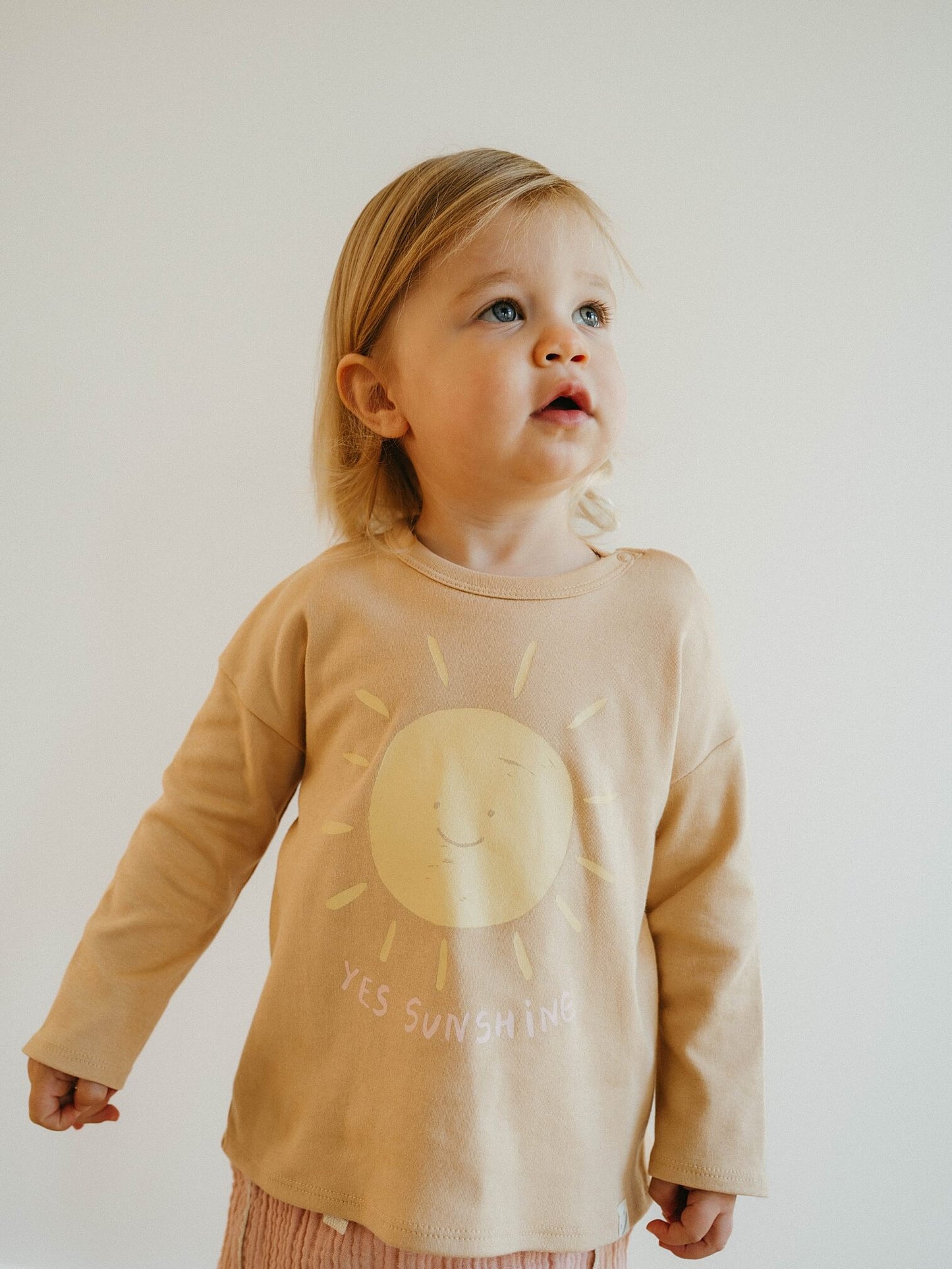 Child wearing a beige long-sleeve shirt with a sun design and text on a plain background