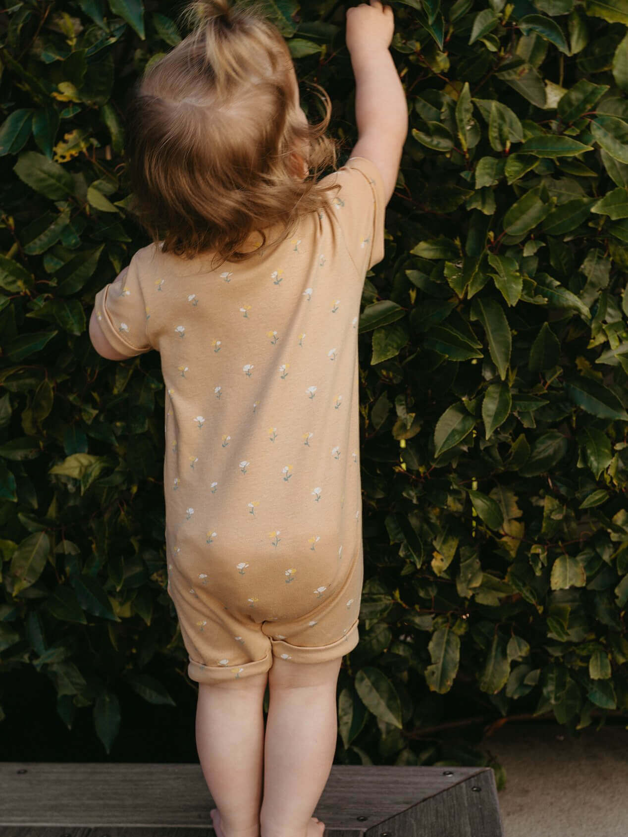 Child in a beige outfit with white flowers standing against a green hedge.
