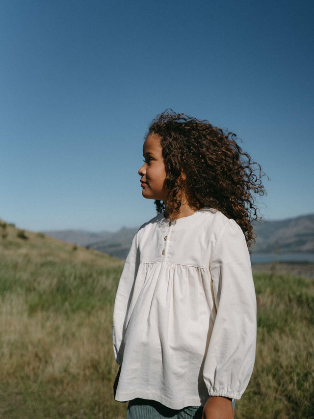 Young girl in a white blouse standing in a grassy field with mountains in the background