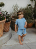 A young child wearing a light blue short-sleeve t-shirt and matching shorts walks barefoot on a concrete patio next to large potted plants.