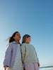 Two young girls standing side-by-side on a beach under a clear blue sky. The girl on the left wears a light purple button-up shirt over a white and blue striped dress, and the girl on the right wears a pale green textured knit sweater with a pink skirt.