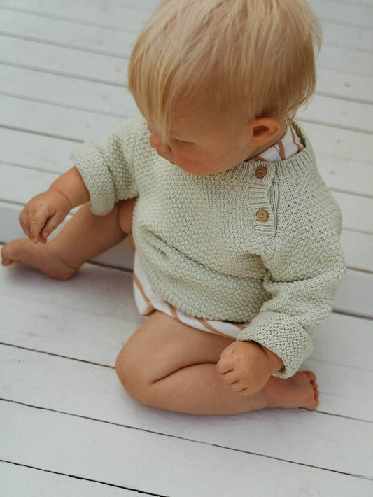 Baby wearing a green knitted sweater sitting on a wooden floor