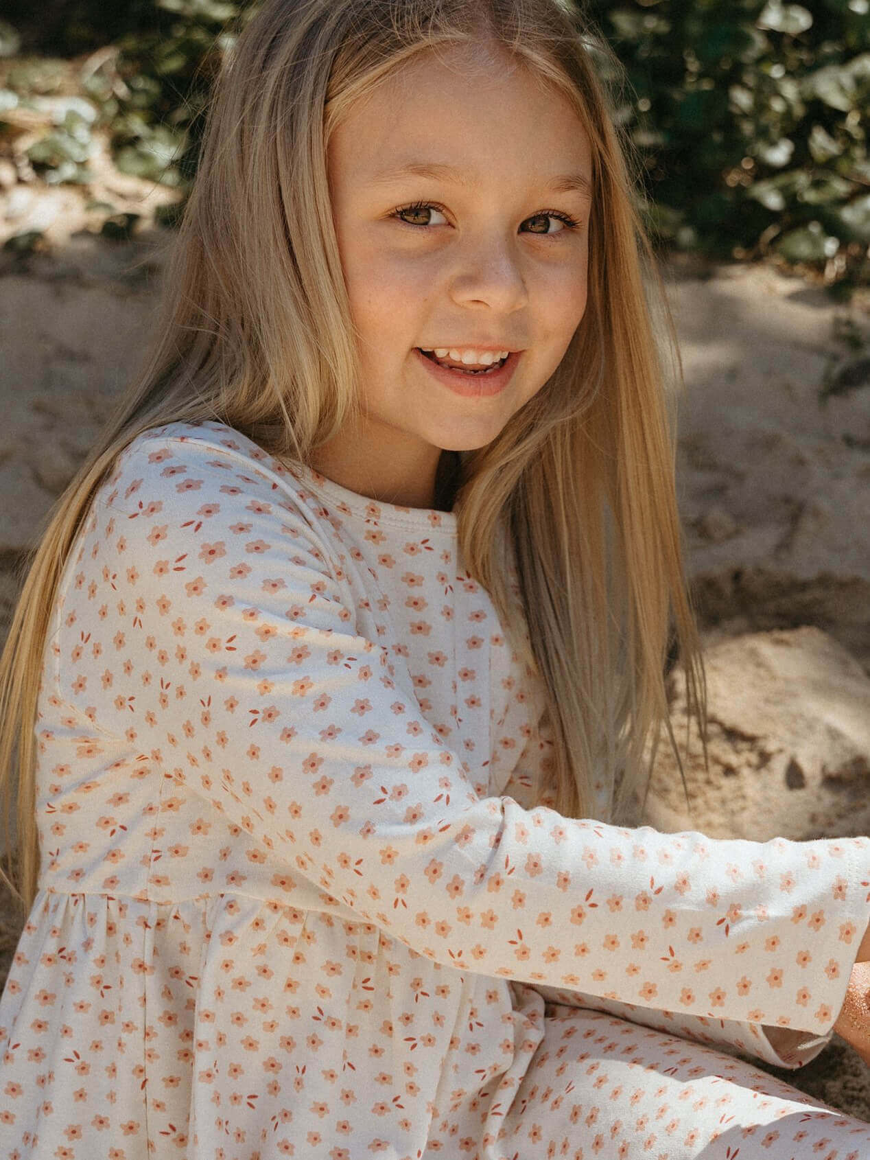 Young girl with long blonde hair wearing a white dress with small patterns, sitting outdoors.