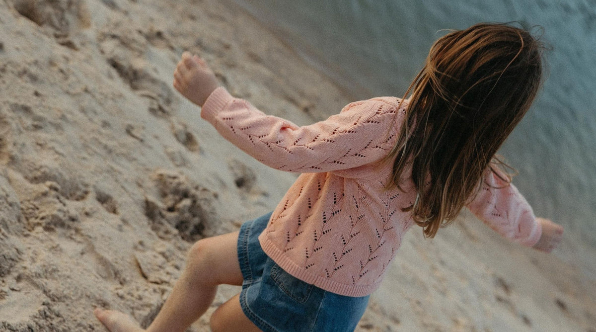 A young girl with long brown hair is seen from behind, wearing a light pink knit sweater and denim shorts while sliding down a sandy hill.